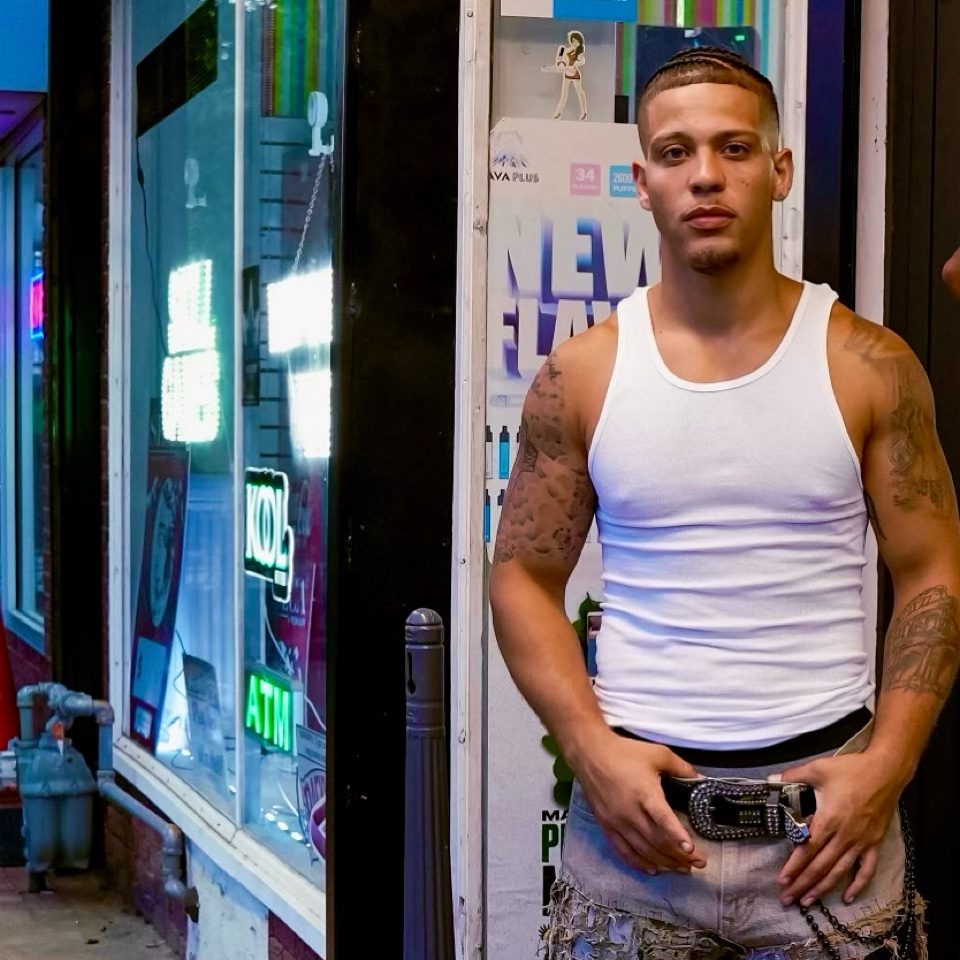 Rome Rizzo modeling a white tank top and jeans outside a neon-lit storefront at night