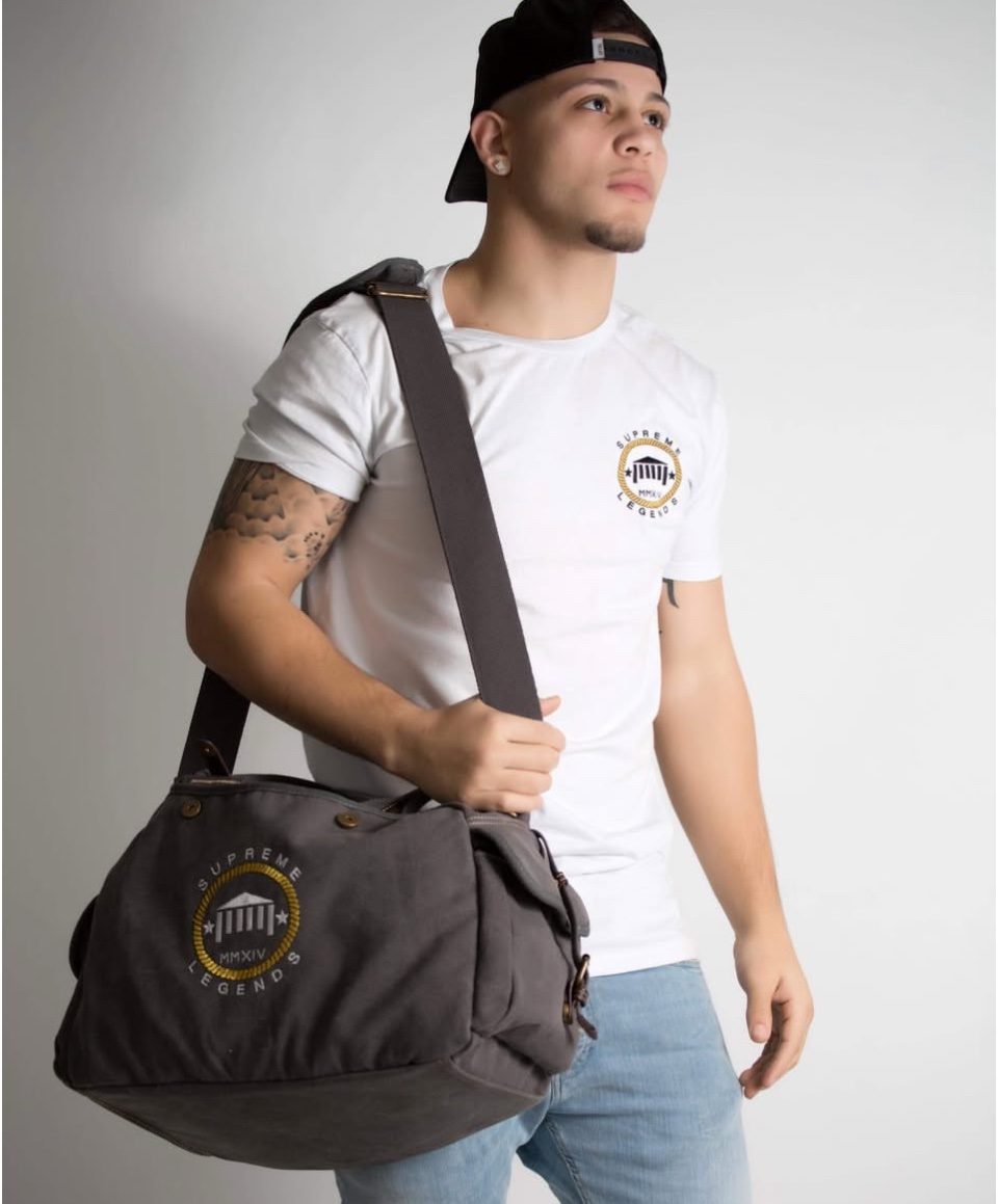 Rome Rizzo in a white T-shirt and backwards cap holding a duffel bag in a casual studio portrait